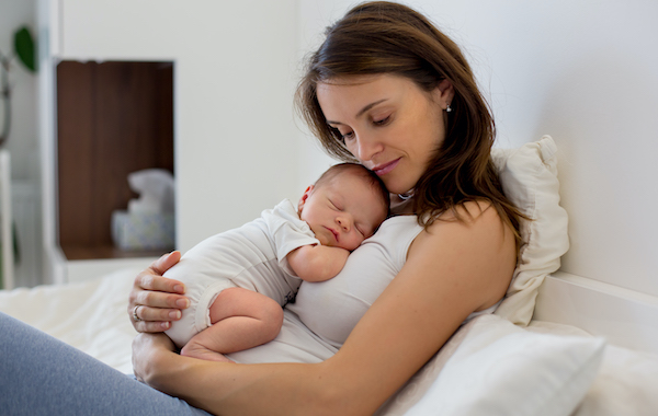 Young mother lying in bed with her newborn baby boy, holding him in her arms and smiling from happiness